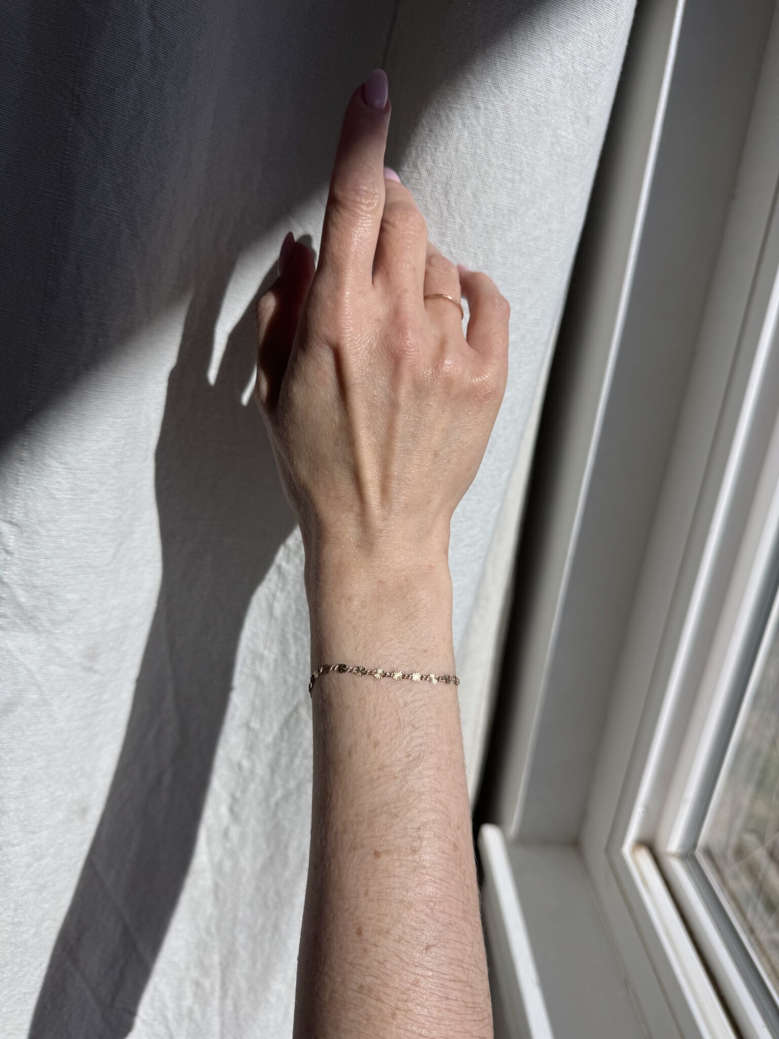 A hand with a gold chain bracelet is raised near a sunlit window, casting a shadow on a light-colored textured wall.