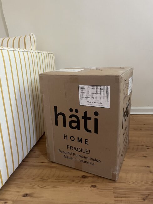 A cardboard box labeled “hati HOME” sits on a wooden floor next to a striped sofa. The box is marked “FRAGILE” and “Made in Indonesia.”.