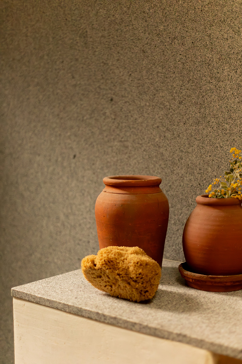 Two clay pots and a natural sponge rest on a gray countertop against a textured gray wall; one pot holds dried yellow flowers.