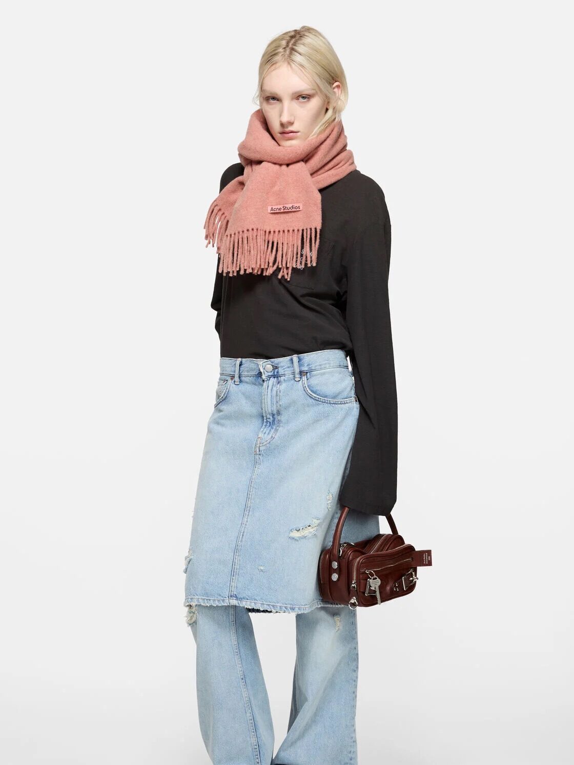 Person standing against a white background wearing a pink scarf, black long-sleeve top, distressed denim skirt over jeans, black sandals, and holding a small brown handbag.