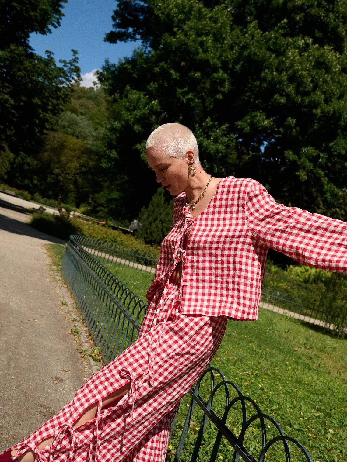 A person with a shaved head wearing a red and white checkered outfit stands by a black metal fence in a park on a sunny day.