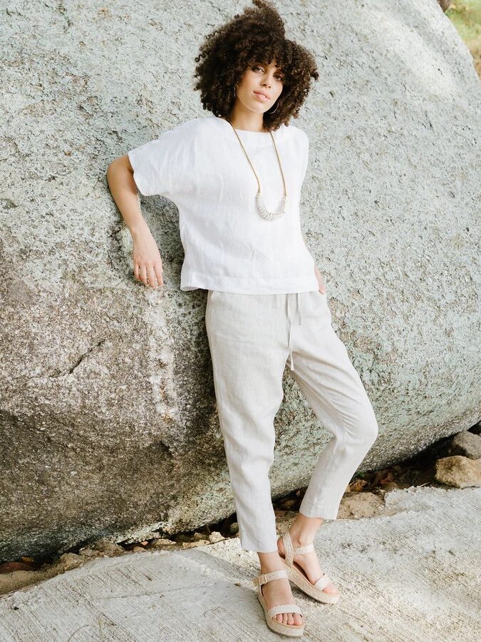 A person with curly hair stands outdoors against a large rock, wearing a white short-sleeve shirt, light-colored pants, sandals, and a long necklace.