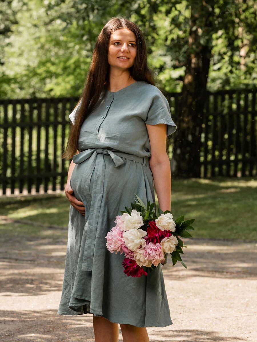 A pregnant woman in a light green dress stands outdoors holding a bouquet of pink and white flowers, with trees and a black wooden fence in the background.