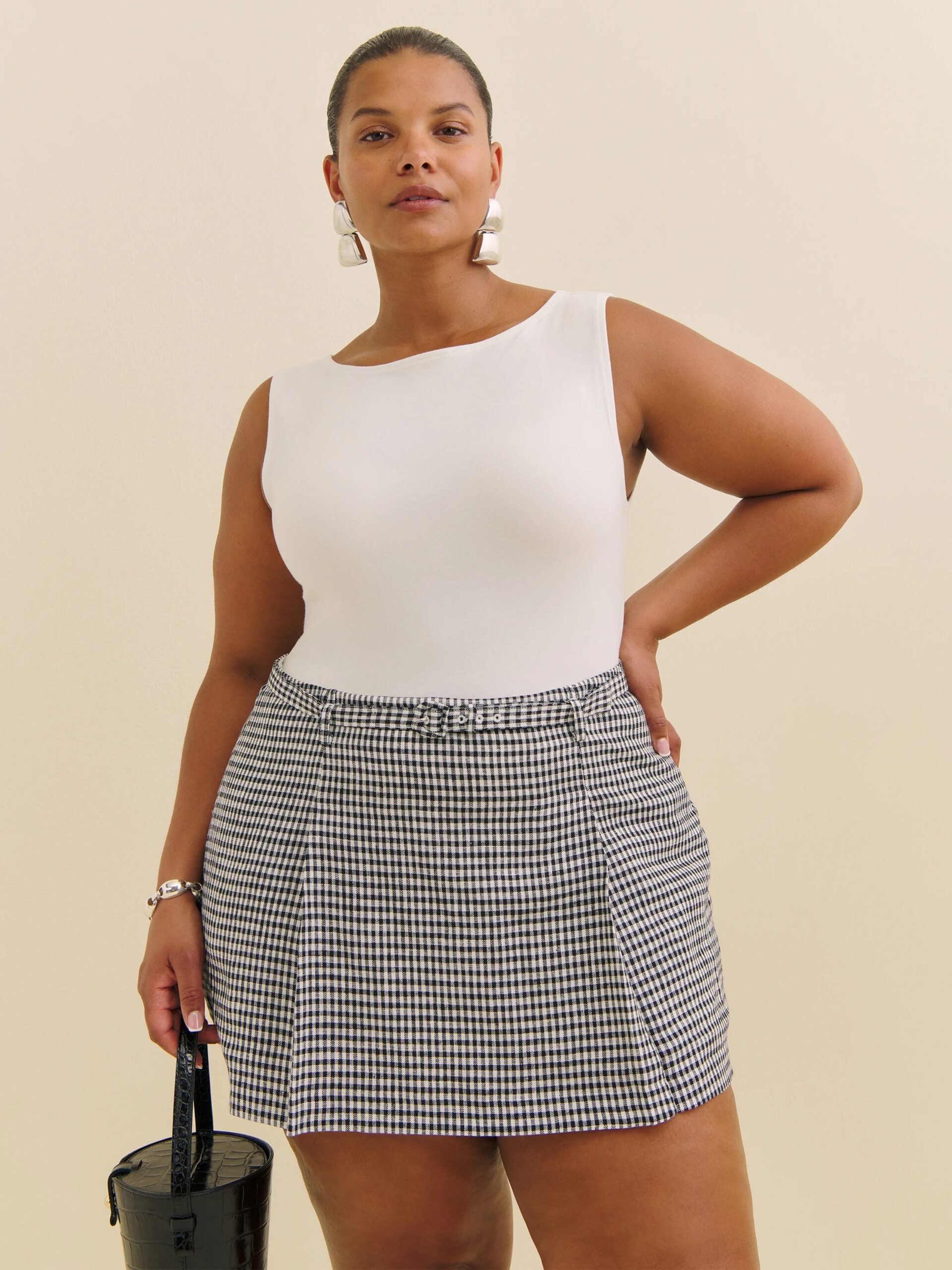 A woman wearing a white sleeveless top and a high-waisted black-and-white checked skirt stands against a plain background, holding a black handbag.