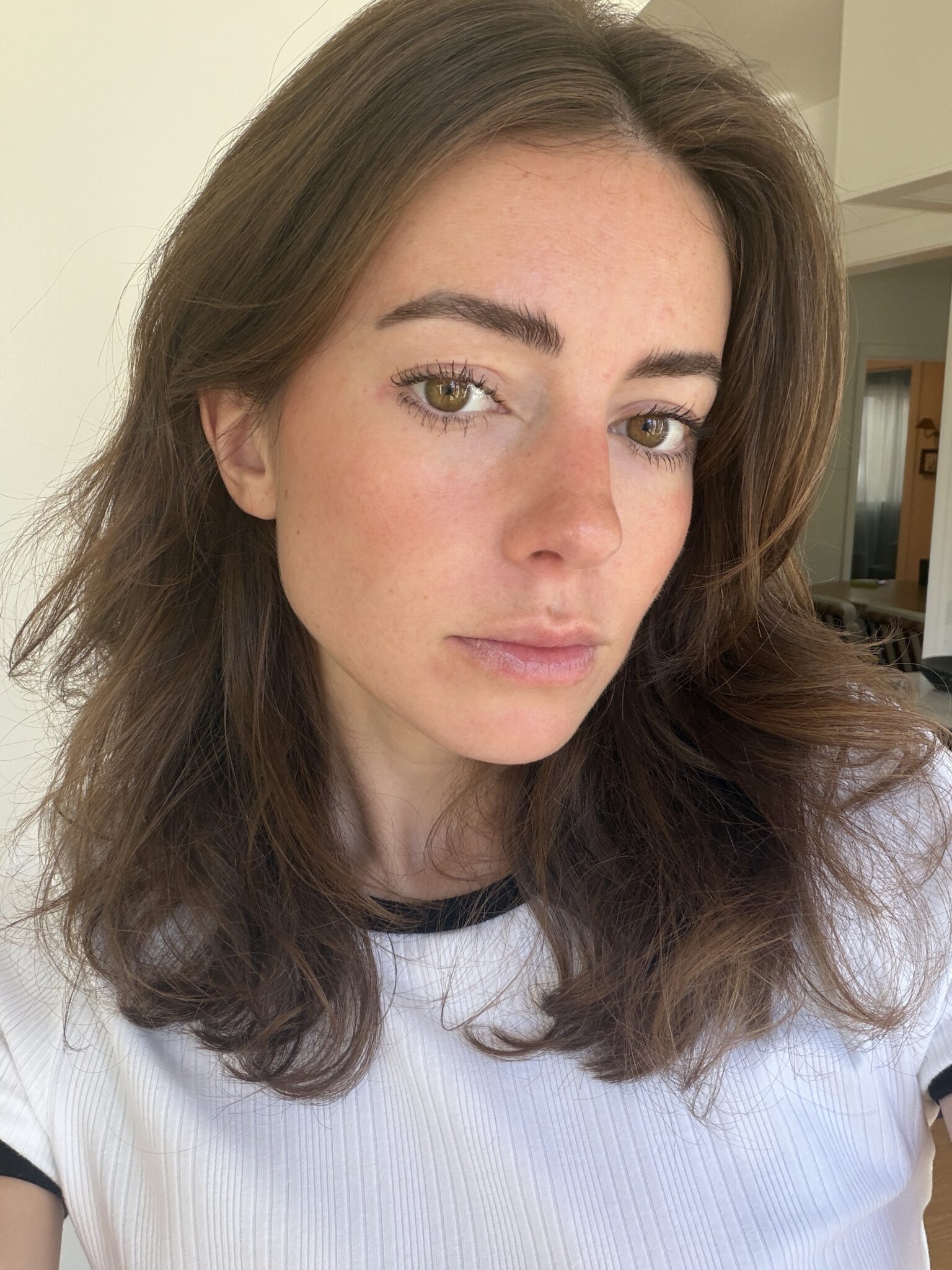 A woman with long brown hair and a white shirt looks directly at the camera; she is indoors with natural light illuminating her face.