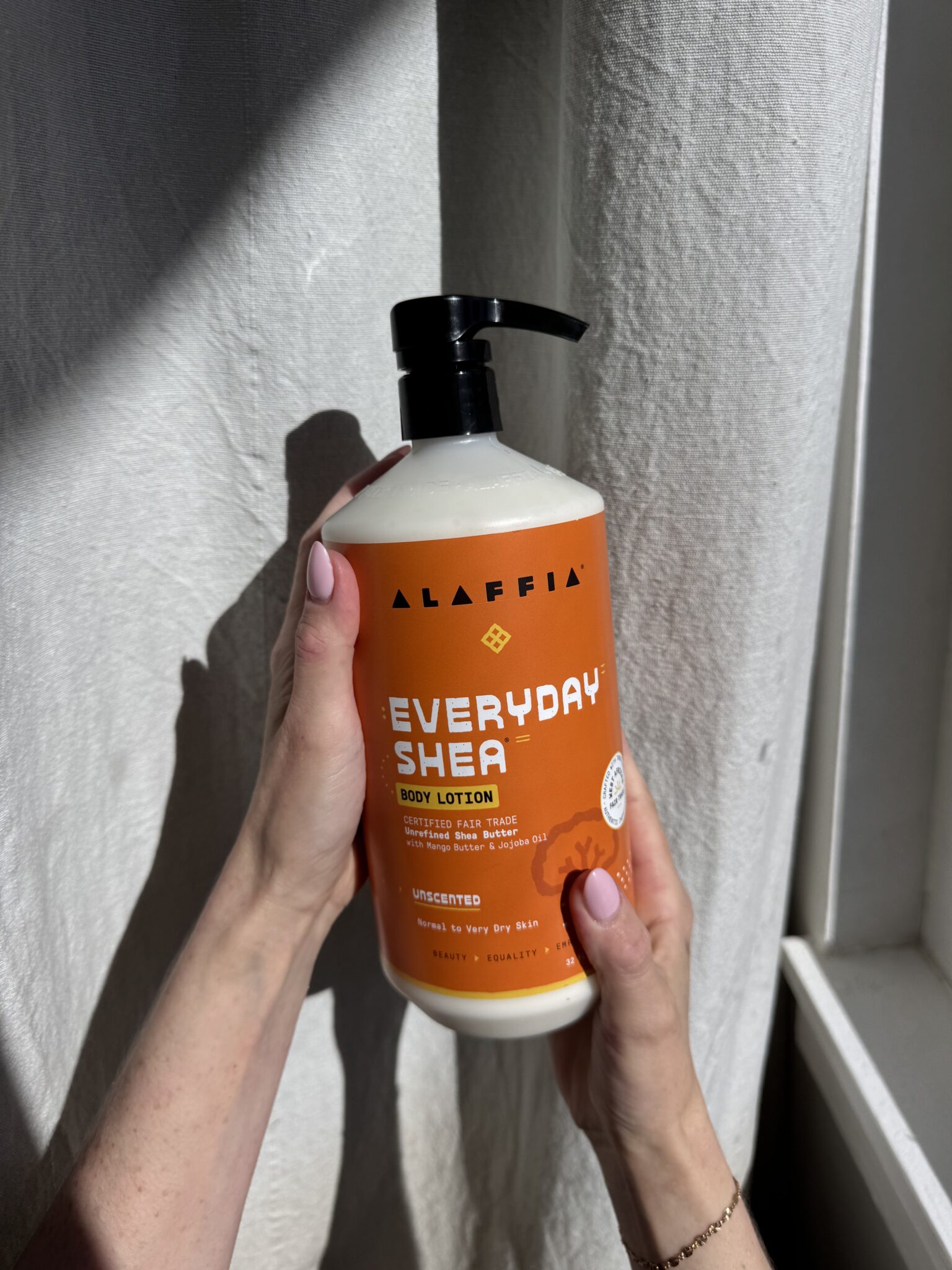 A person holds a large bottle of Alaffia Everyday Shea unscented body lotion with both hands near a sunlit white curtain.