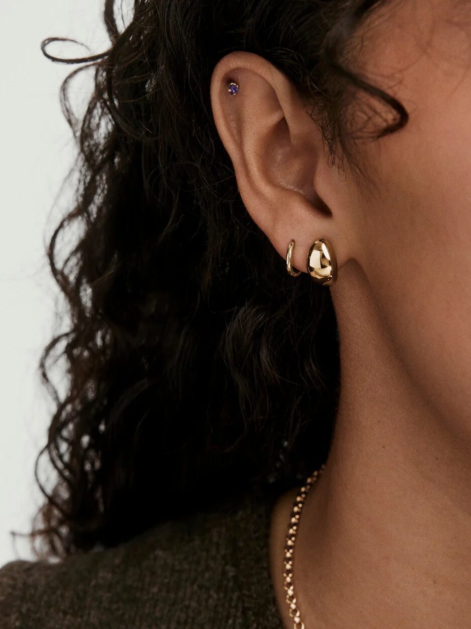 Close-up of a person's ear with three earrings: a small purple stud in the upper ear, a small gold hoop, and a chunky gold earring in the lobe; curly hair and a gold necklace are visible.