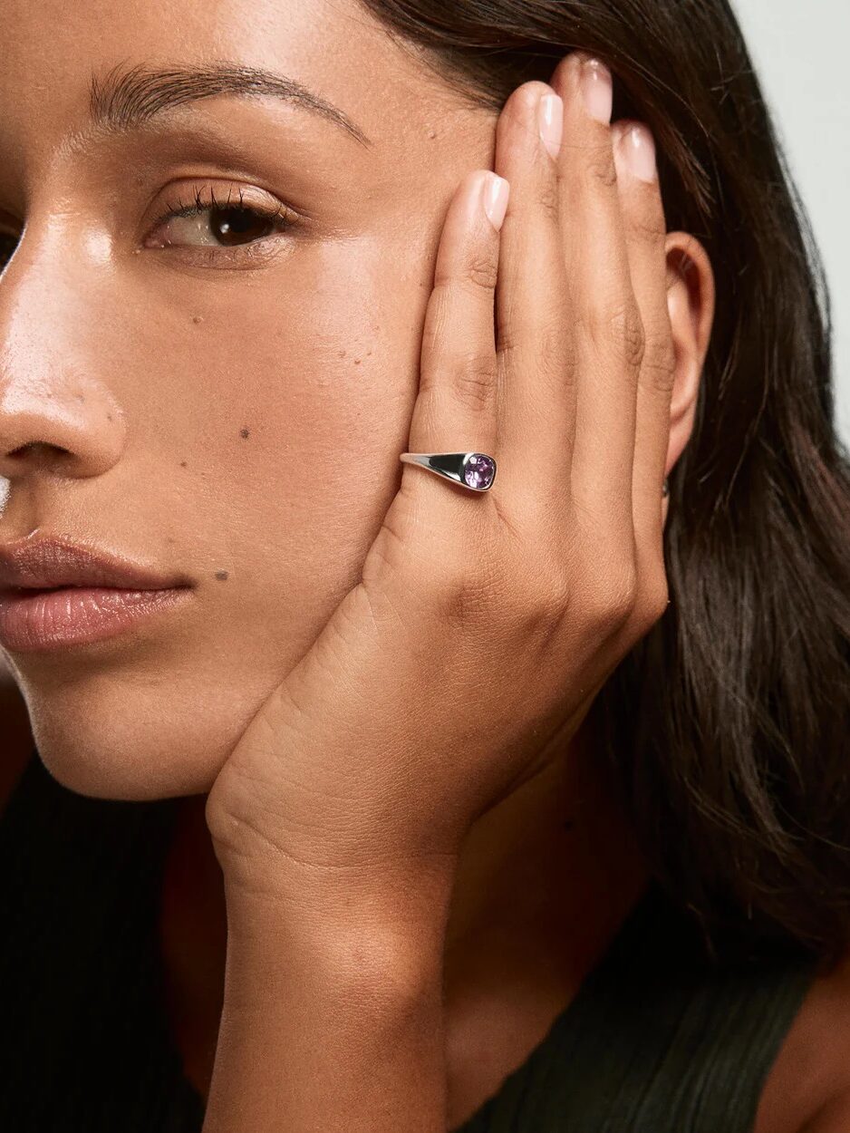 A woman with long dark hair rests her hand on her face, showing a silver ring with a purple gemstone on her finger.