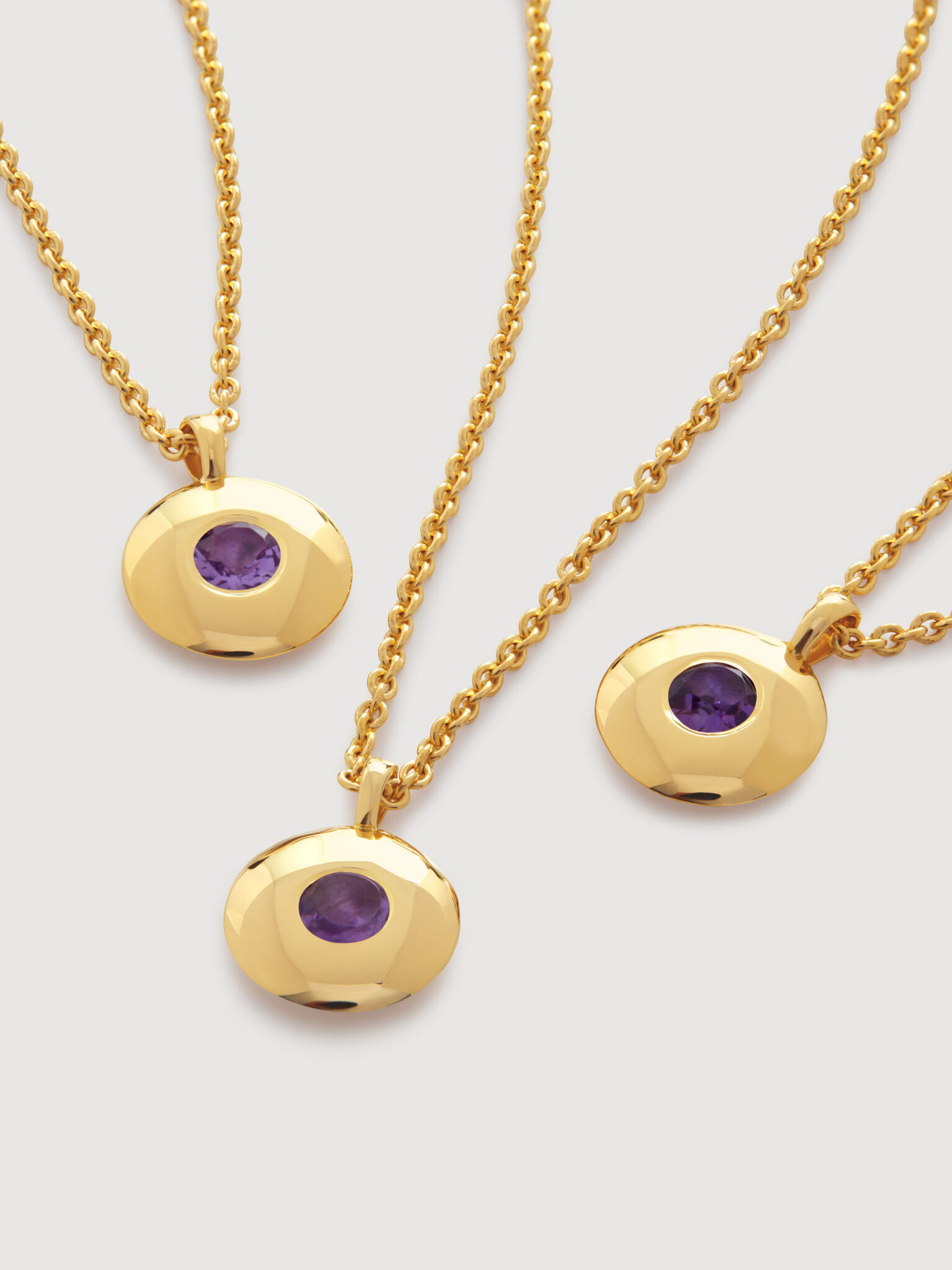 Three gold necklaces with round pendants featuring purple gemstones are displayed on a light background.