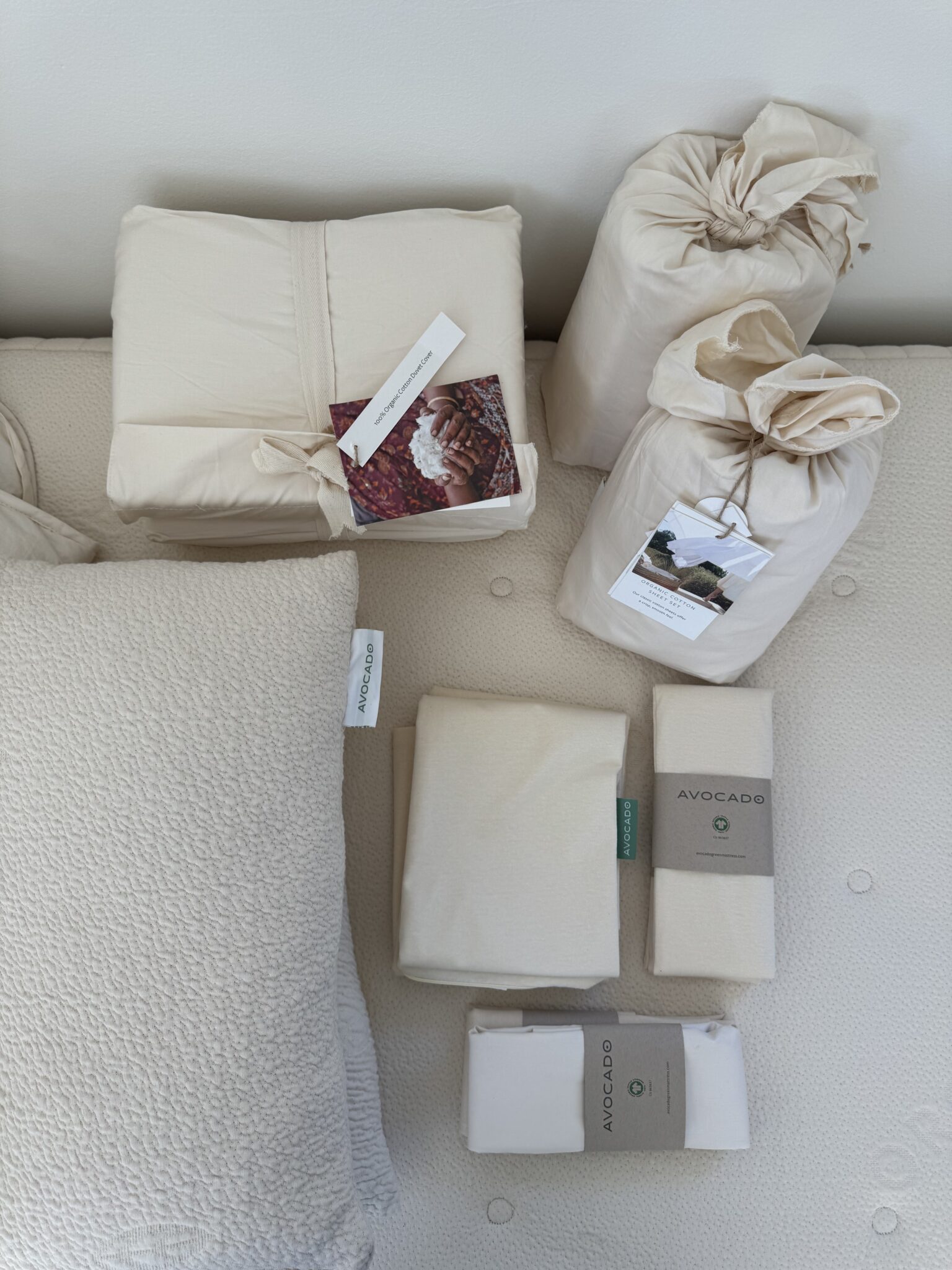 Stacked bedding items, including a pillow, sheets, and fabric bundles, are arranged on a mattress. Each item has a label or tag, and the colors are mostly off-white or beige.