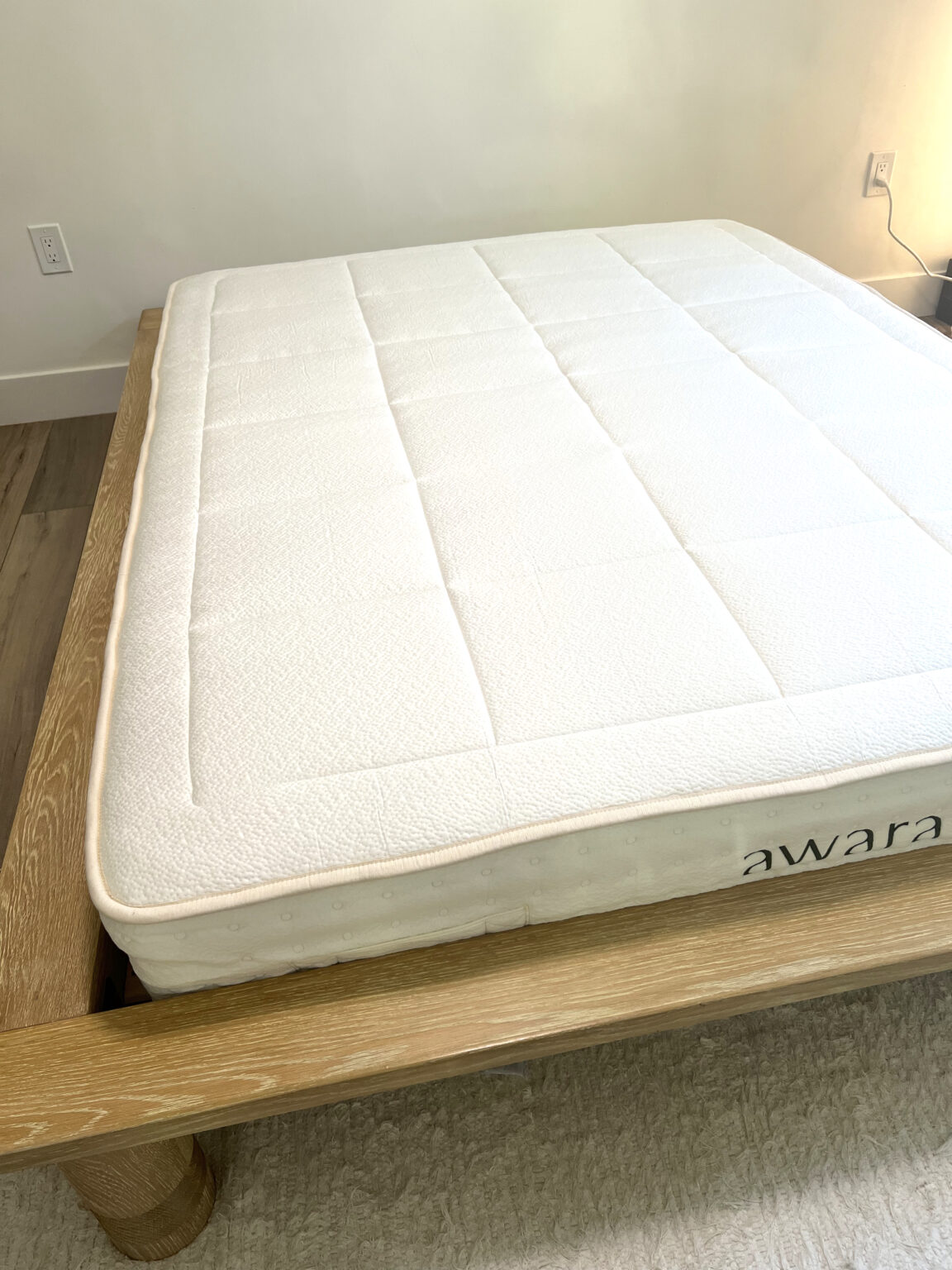A neatly made bed with a white organic mattress labeled "awara" on a light wooden frame, placed on a beige rug near a white wall with electrical outlets.