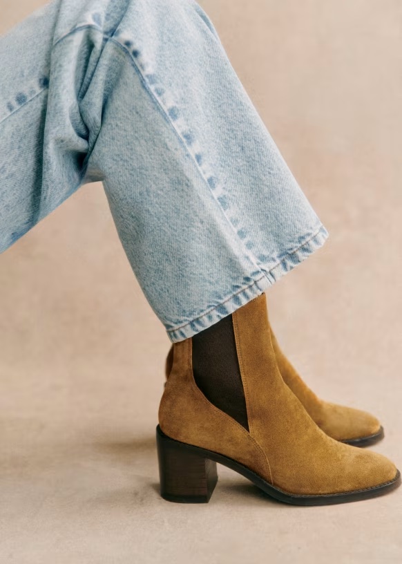 Person wearing light blue jeans and tan suede heeled ankle boots with dark elastic side panels, seated against a neutral background.