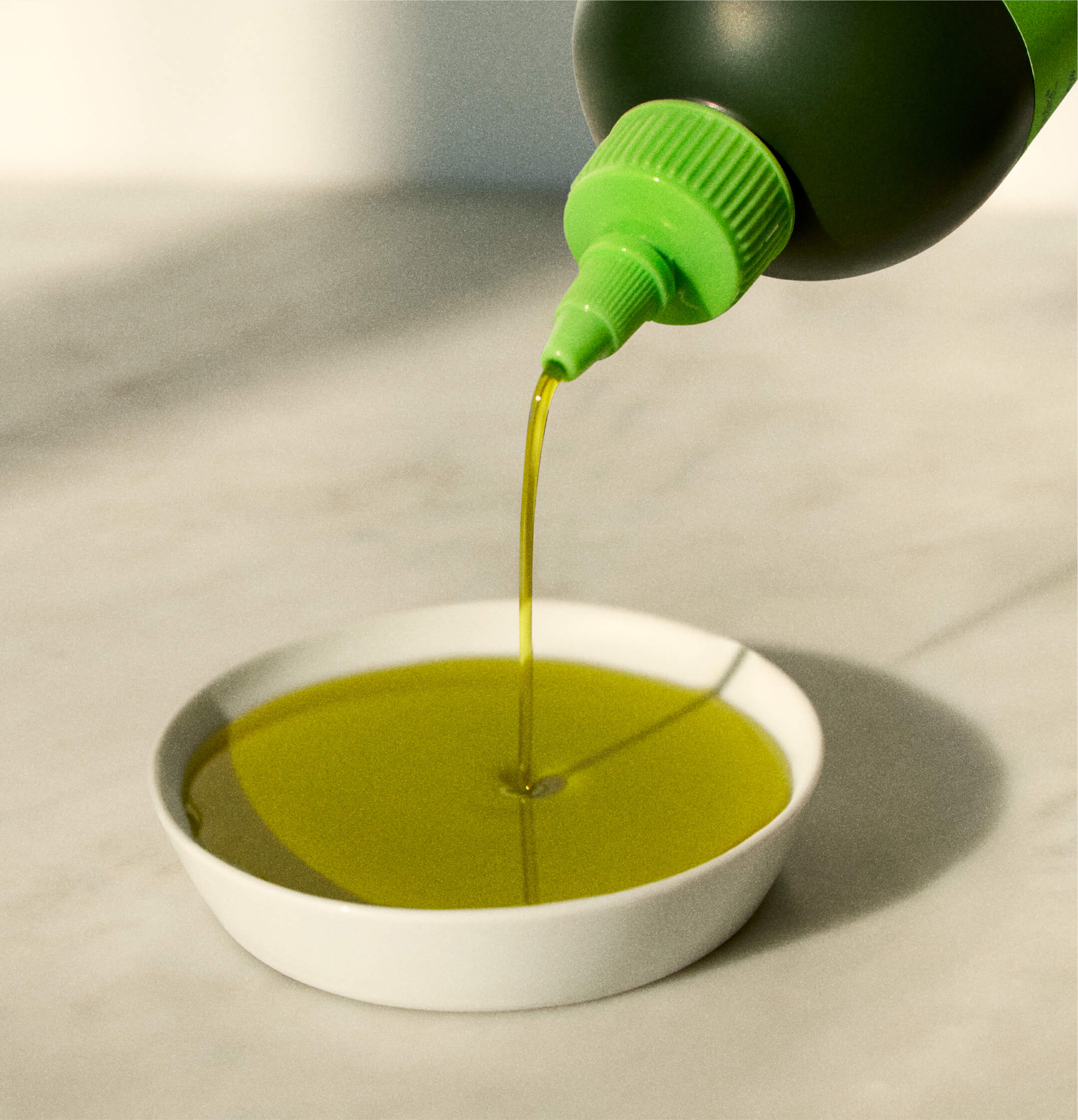 Green oil bottle pouring yellow-green liquid into a small white dish on a light surface, casting soft shadows.