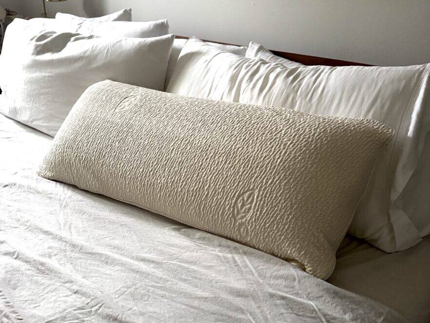 A neatly made bed with white sheets atop an organic mattress, three pillows against the headboard, and a long textured beige body pillow in front.