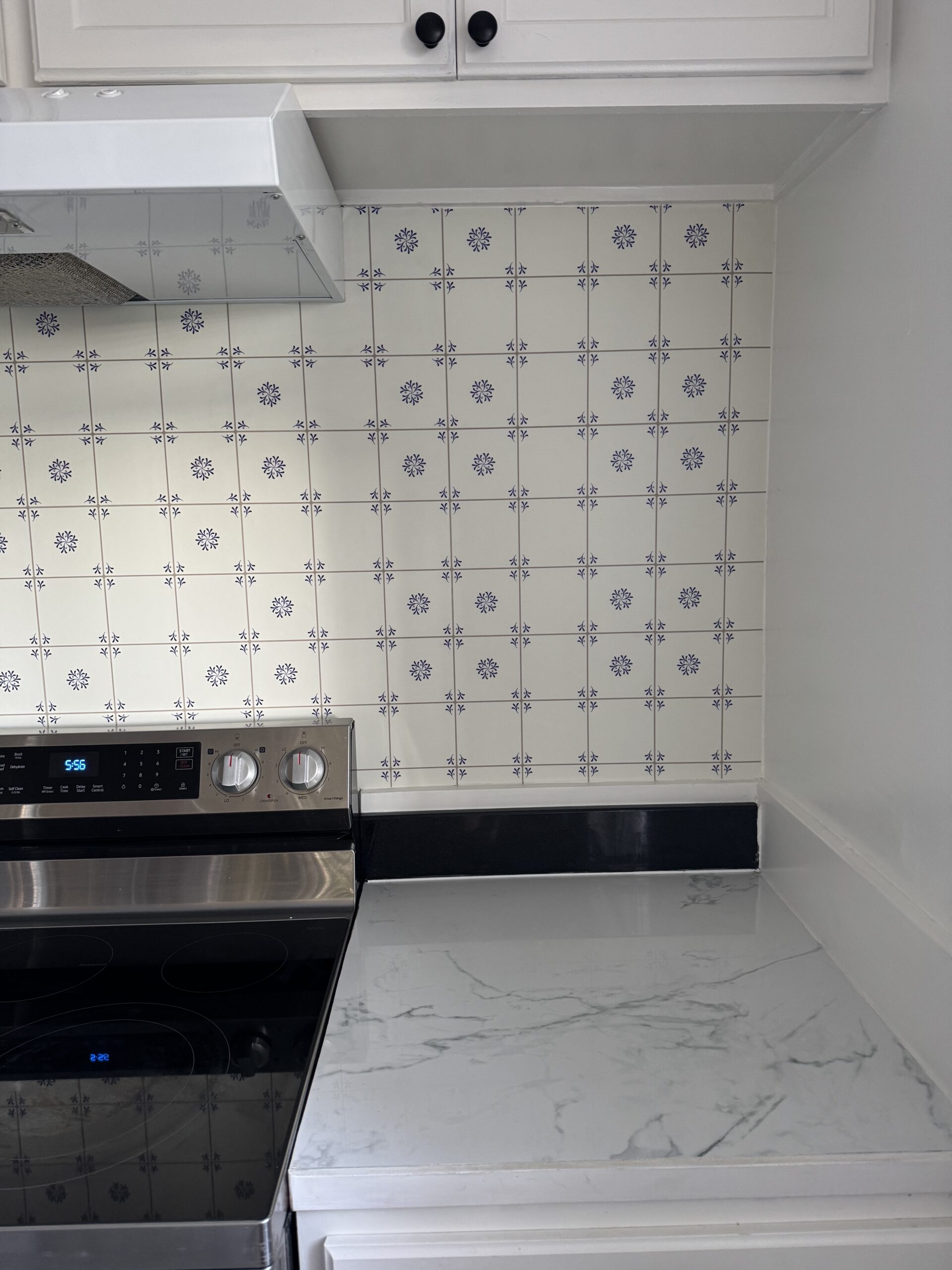 A kitchen corner with a marble countertop, a stainless steel stove, white cabinets, and a tiled backsplash with a blue floral pattern.
