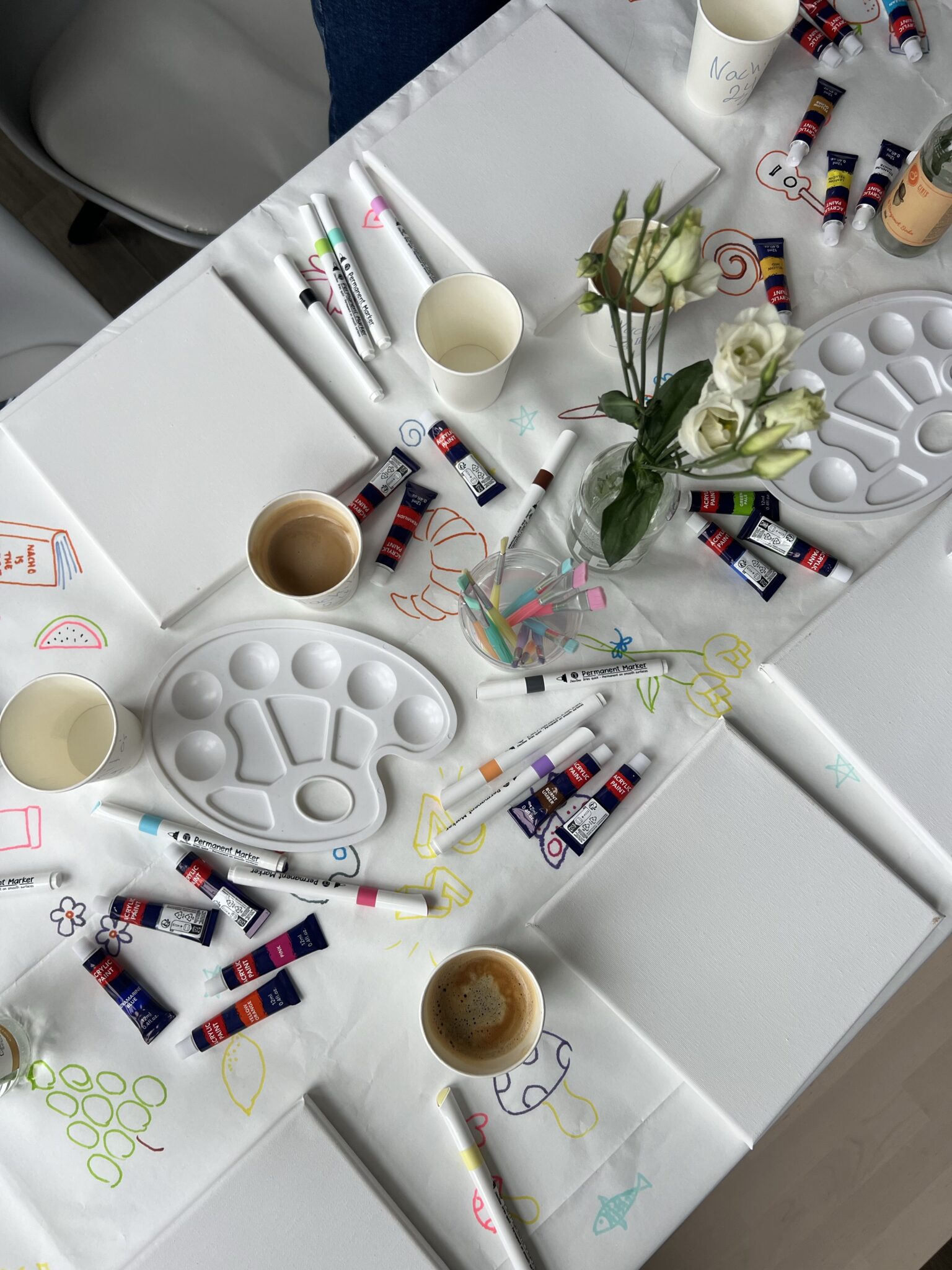Overhead view of a table set up for painting, with canvases, paint tubes, palettes, coffee cups, drawing markers, and a vase of white roses on a decorated tablecloth.