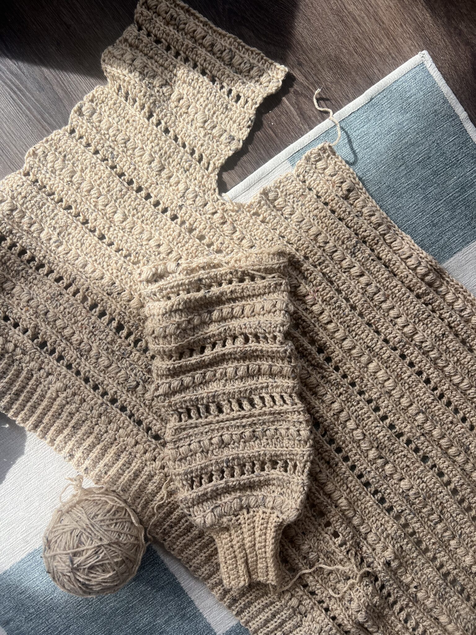 A partially completed crocheted garment and a finished sleeve rest on a mat, with a ball of yarn nearby in sunlight.