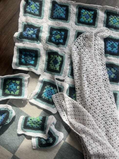 A partially completed crocheted blanket with blue and green square patterns, several loose squares, and a separate piece of white crochet fabric lying on a checkered surface.