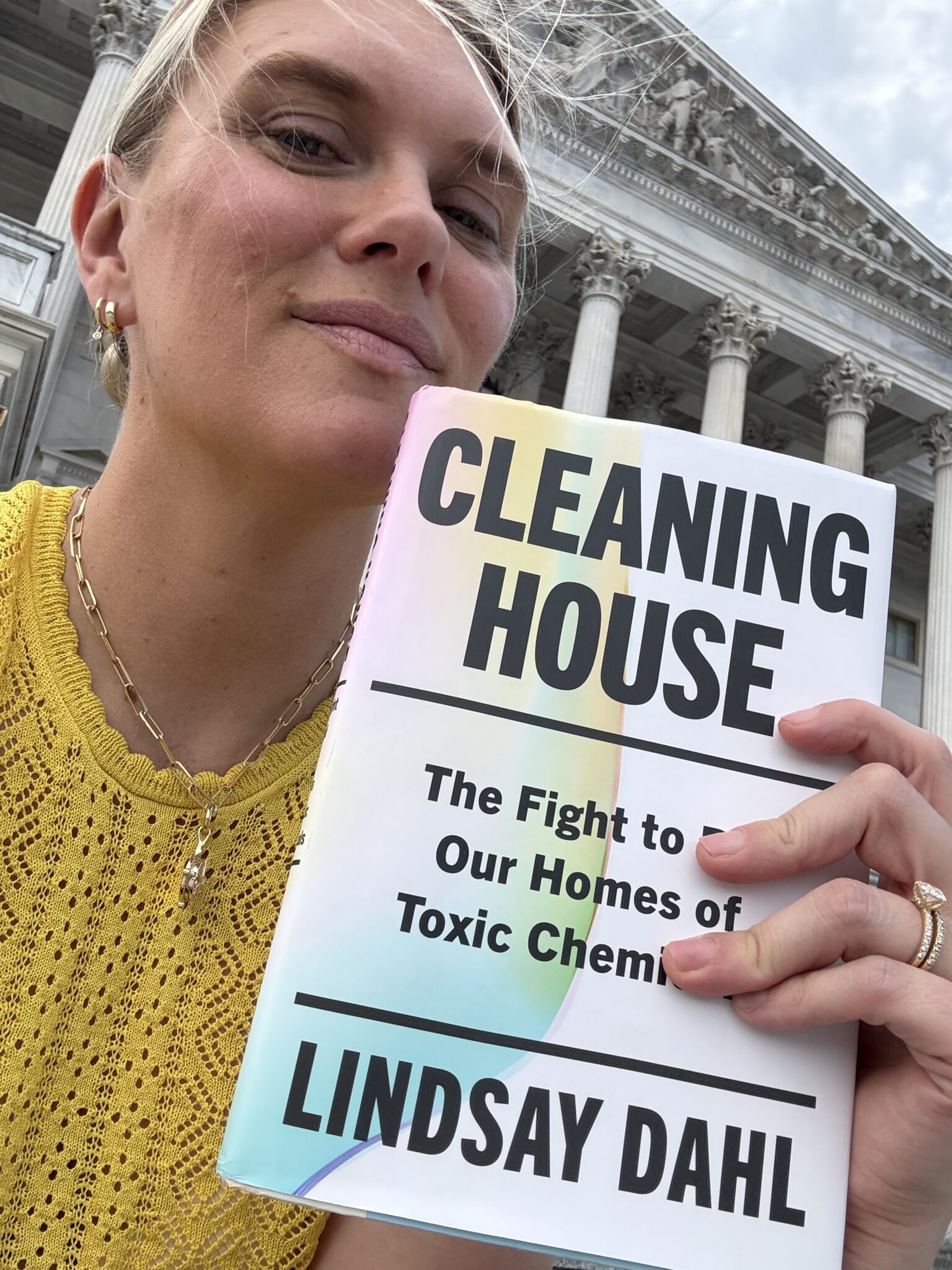 A woman in a yellow top holds up the book "Cleaning House: The Fight to Our Homes of Toxic Chemicals" by Lindsay Dahl in front of a large building with columns.