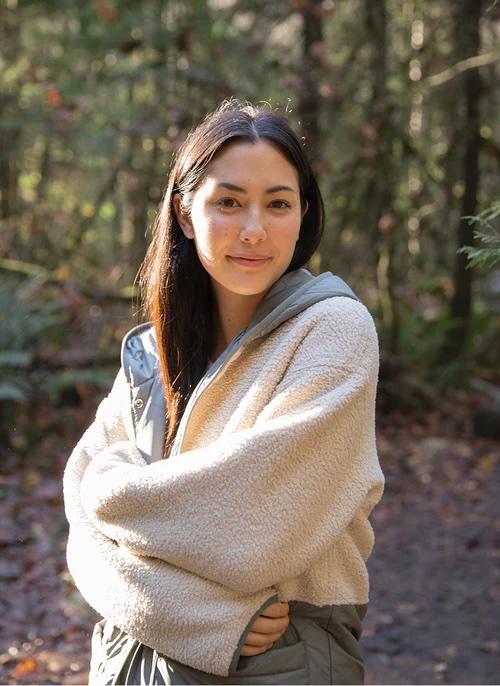 A woman with long dark hair stands outdoors on a forest trail, wearing a beige fleece jacket and looking at the camera with a slight smile.