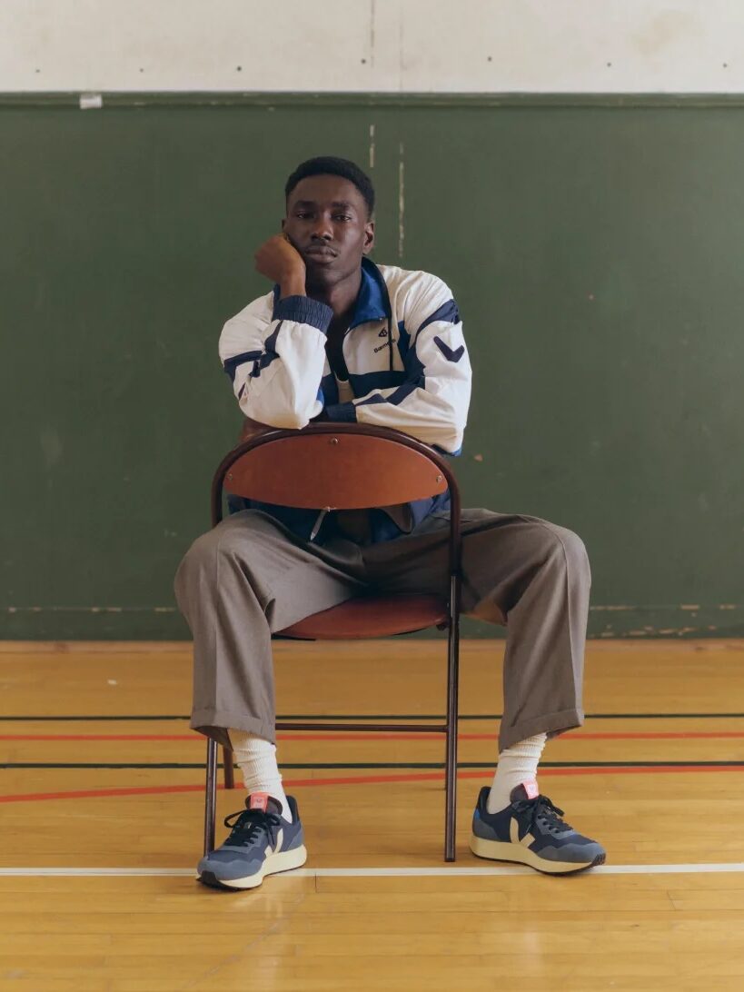 A person sits on a chair in a gymnasium, resting their chin on one hand. They wear a white and navy jacket, gray pants, white socks, and dark sneakers.