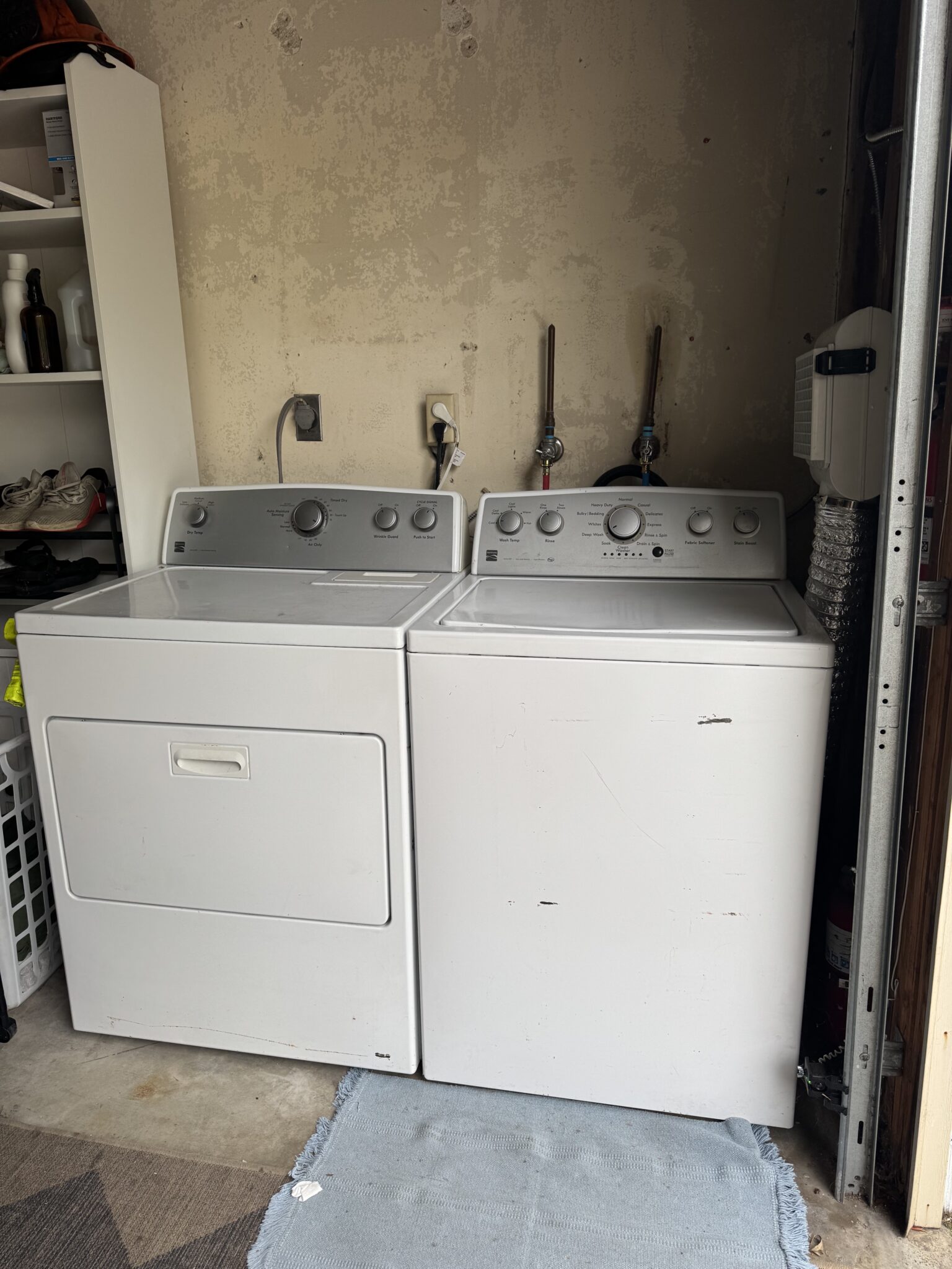 A white dryer and washing machine sit side by side in a laundry area with shelves, laundry baskets, and cleaning supplies nearby.