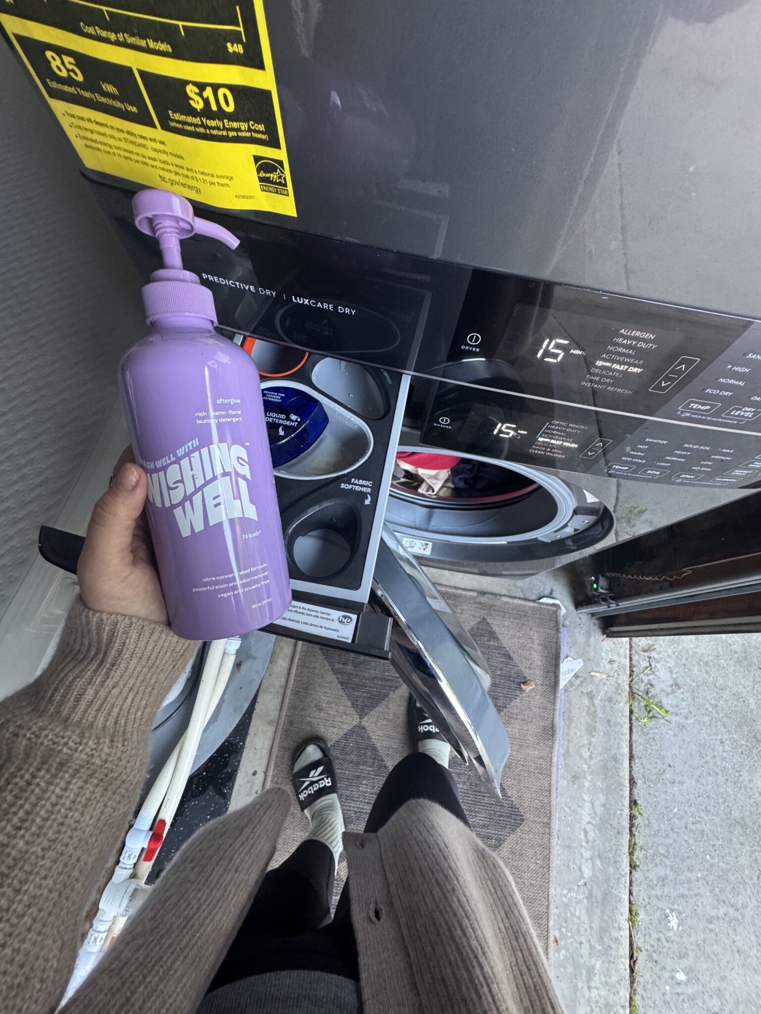 Person holding a purple bottle of Wishing Well laundry detergent in front of an open washing machine with detergent drawer pulled out.