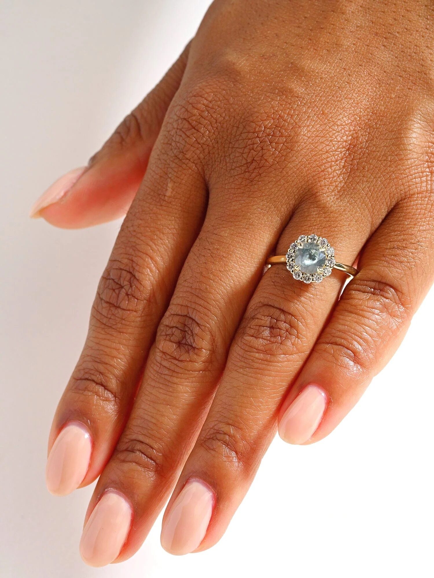 A hand with neatly manicured nails wears a ring featuring a light blue gemstone surrounded by small diamonds on a silver band.