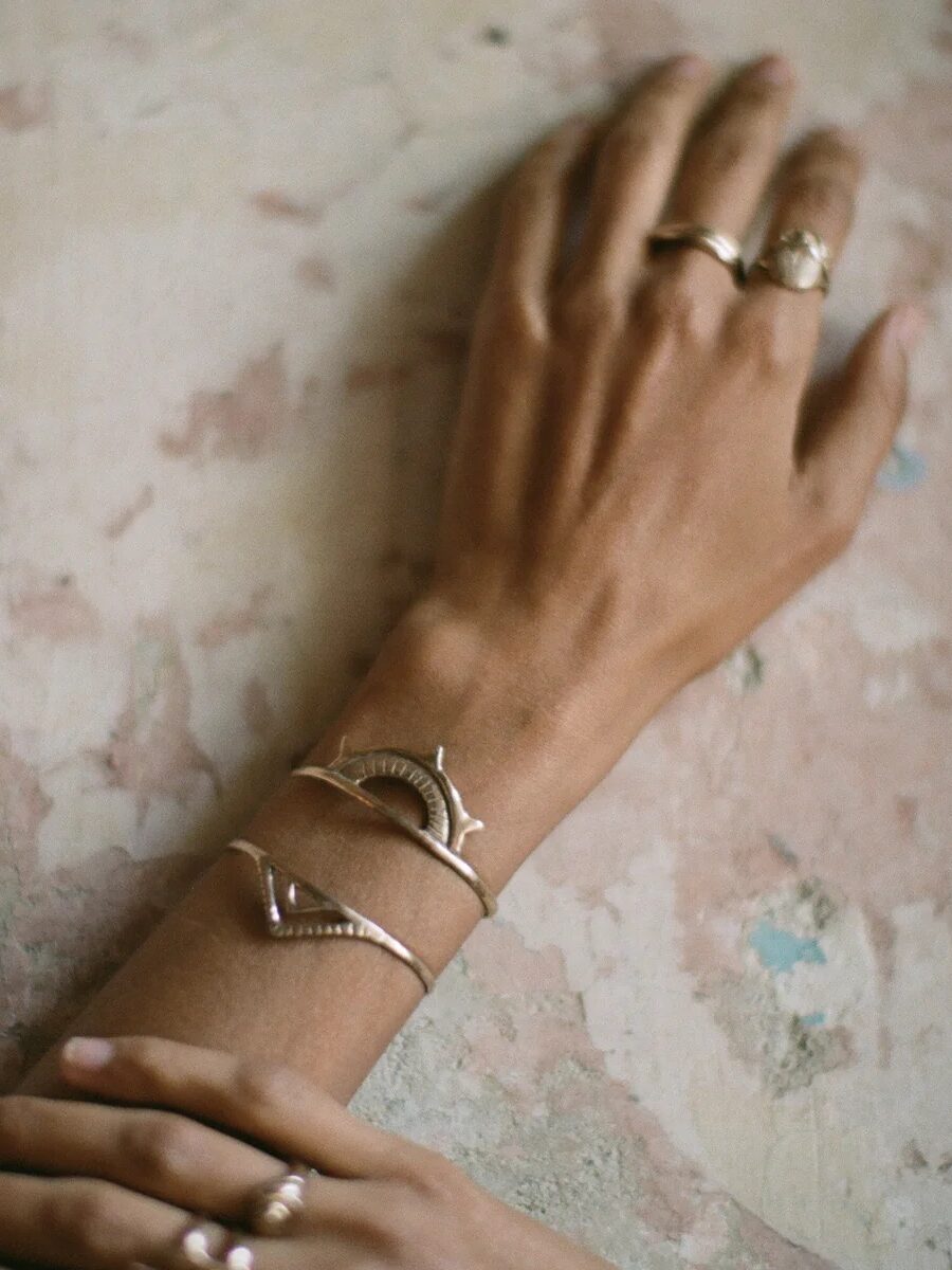 A close-up of a person's hands resting on a textured, worn surface, wearing gold rings and a distinctive gold bracelet shaped like two fish.