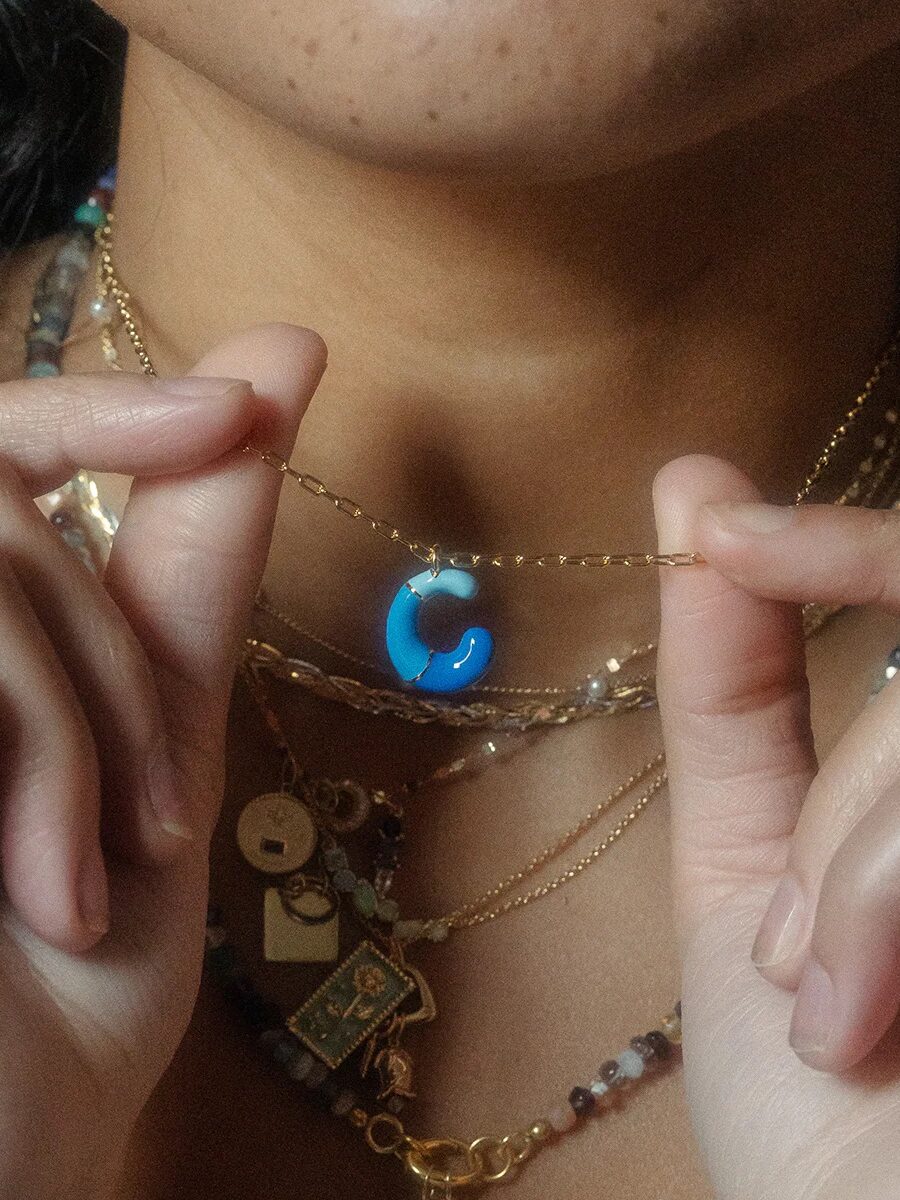 A person holds a gold necklace with a blue and white letter "C" pendant, wearing multiple layered necklaces and rings.