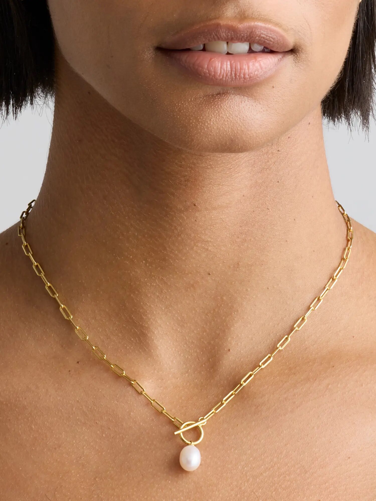 A person wearing a gold chain necklace with a single pearl pendant; only the lower face and neck are visible.