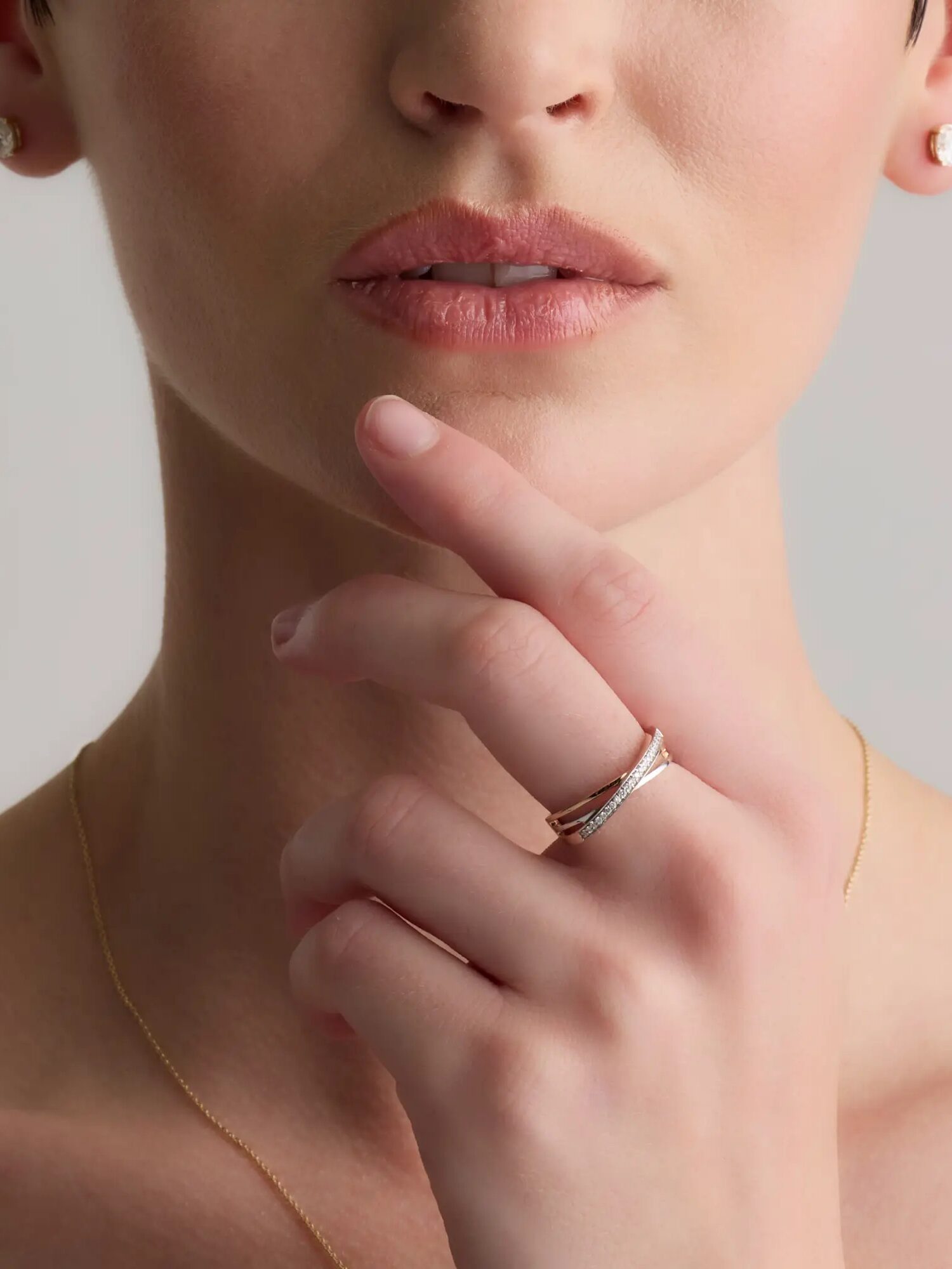 A close-up of a person wearing a gold necklace, stud earrings, and two stacked rings on their hand near their lips.