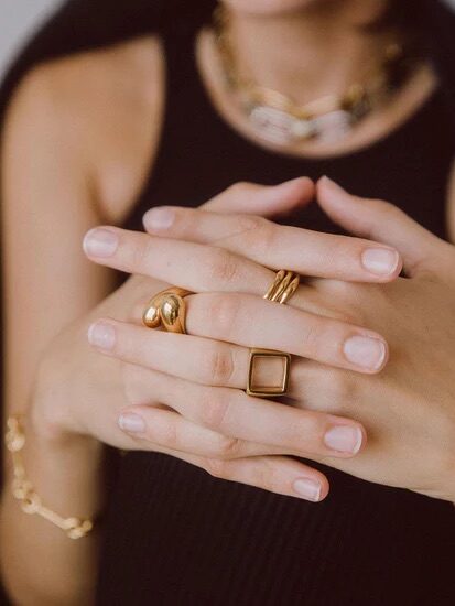 A person wearing multiple gold rings, a bracelet, and a necklace, with hands folded together against a black sleeveless top.
