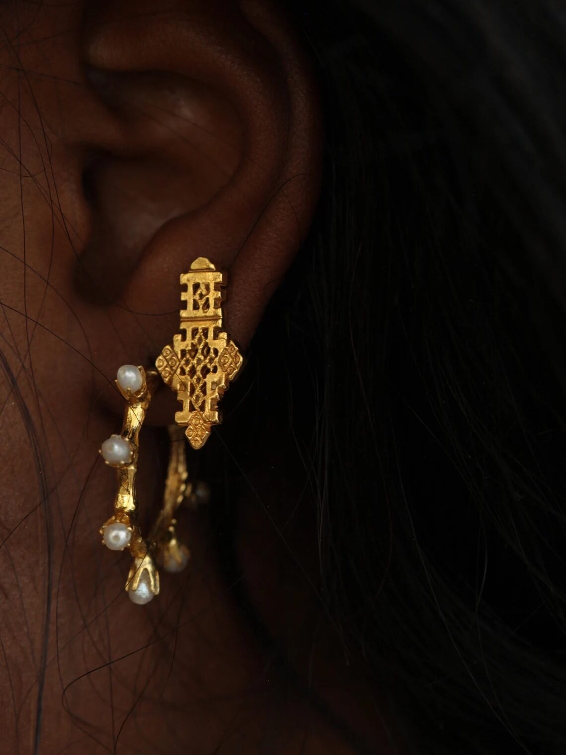 Close-up of a person's ear wearing a gold earring with intricate detailing and small dangling pearls.