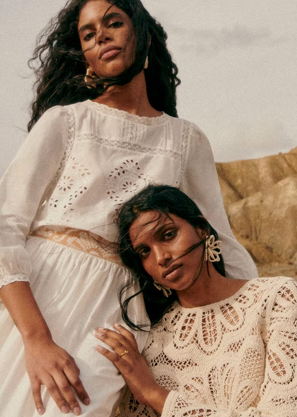 Two women pose outdoors, wearing light-colored, long-sleeved, lace and crochet dresses. One stands while the other leans against her. Wind blows their hair across their faces.