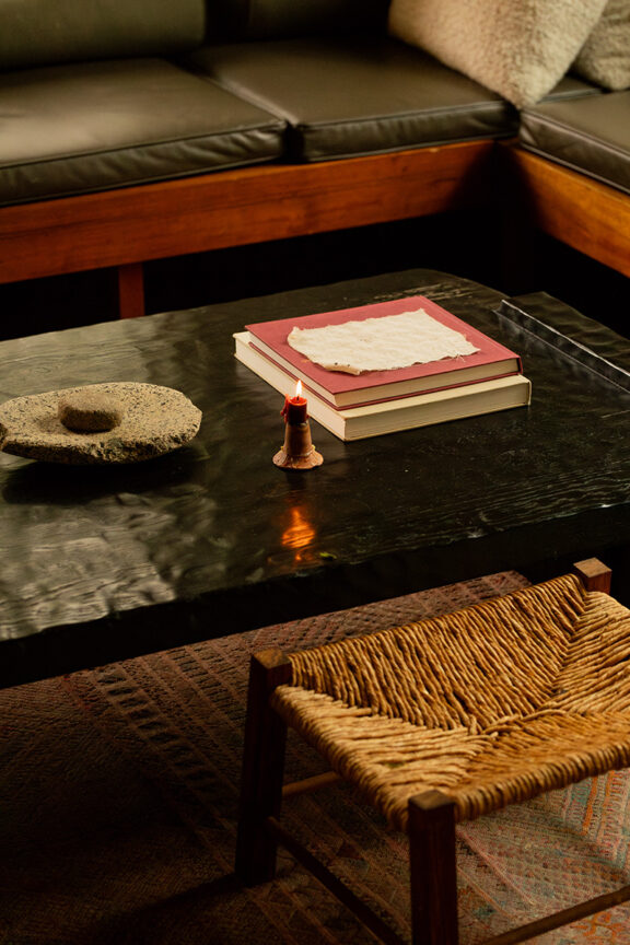 A black metal coffee table with stacked books, a piece of paper, a small lit candle, and a stone object; in front of a woven stool and a black leather sofa.