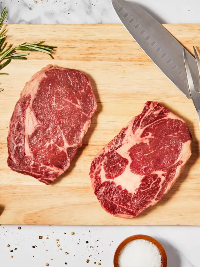 Two raw ribeye steaks on a wooden cutting board with a knife, rosemary, scattered pepper, and a small bowl of salt nearby.