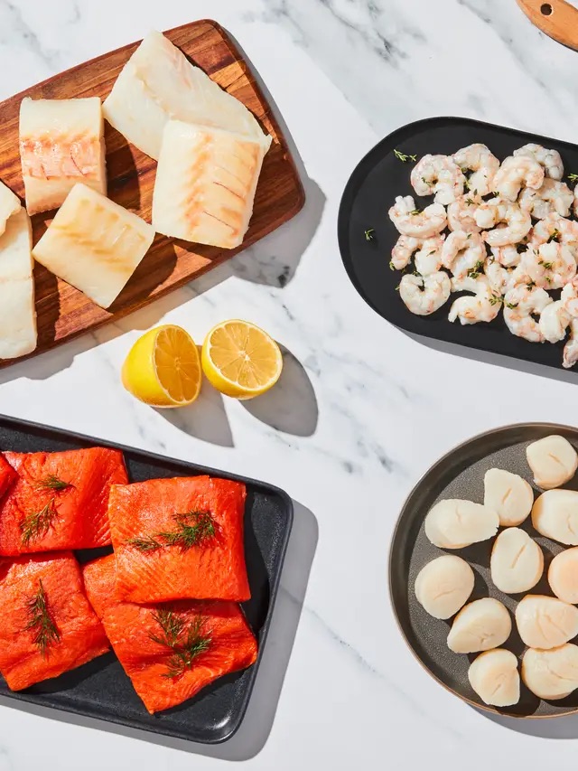 Raw seafood including salmon fillets, white fish pieces, shrimp, and scallops arranged on plates with two lemon halves on a marble surface.