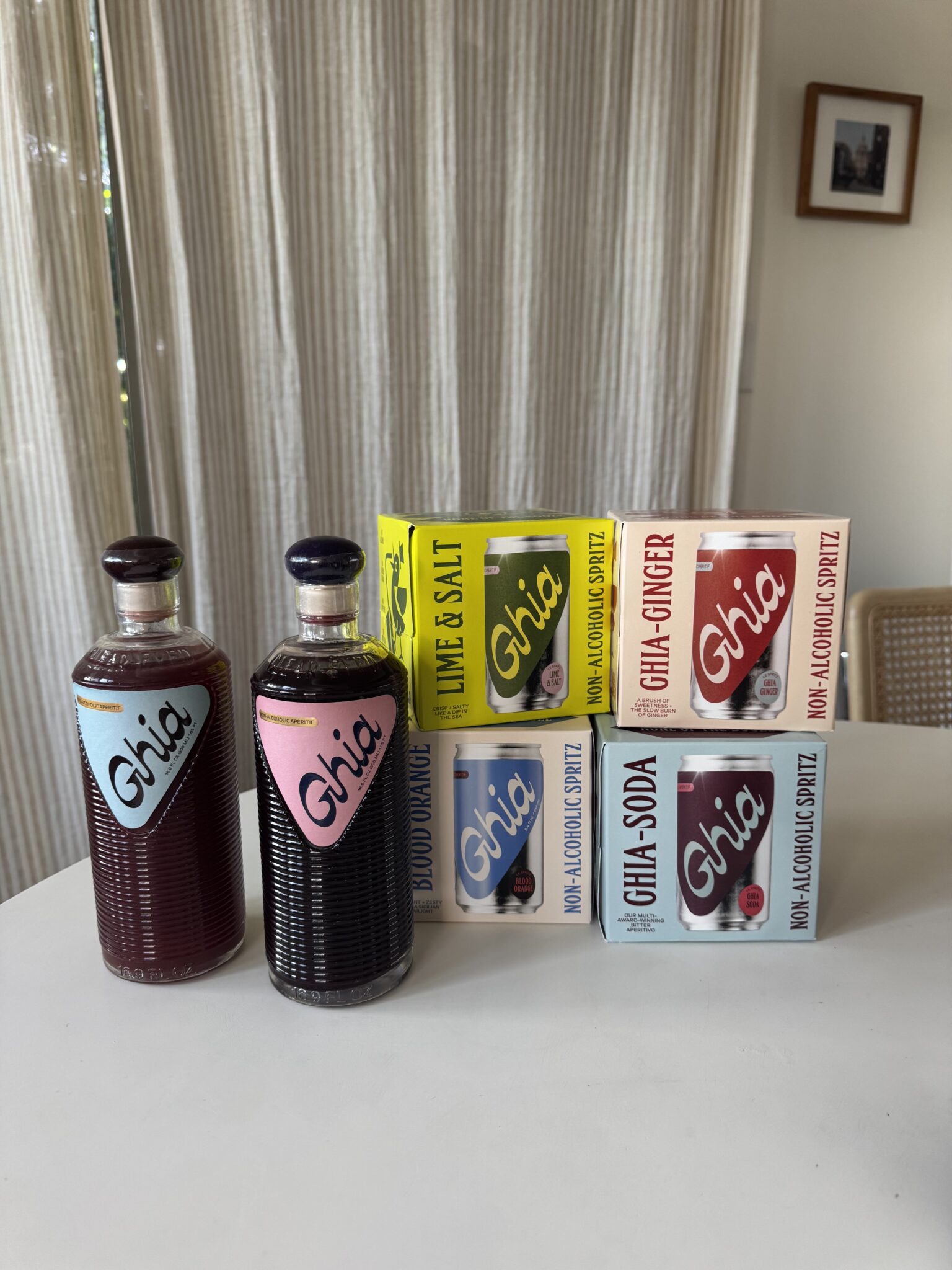 Two glass bottles and four boxes of Ghia non-alcoholic beverages are arranged on a white surface in front of beige curtains.