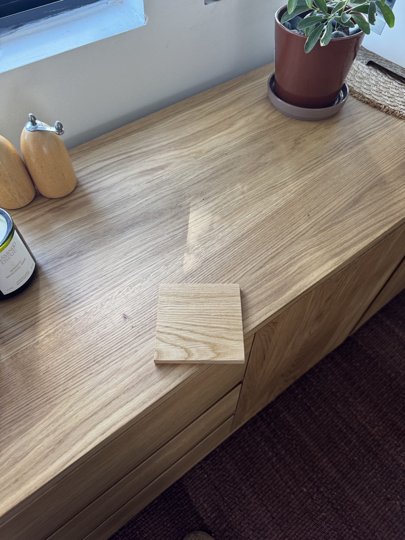 A small wooden board sits on a light wood cabinet next to a potted plant, a candle, and salt and pepper shakers by a window.