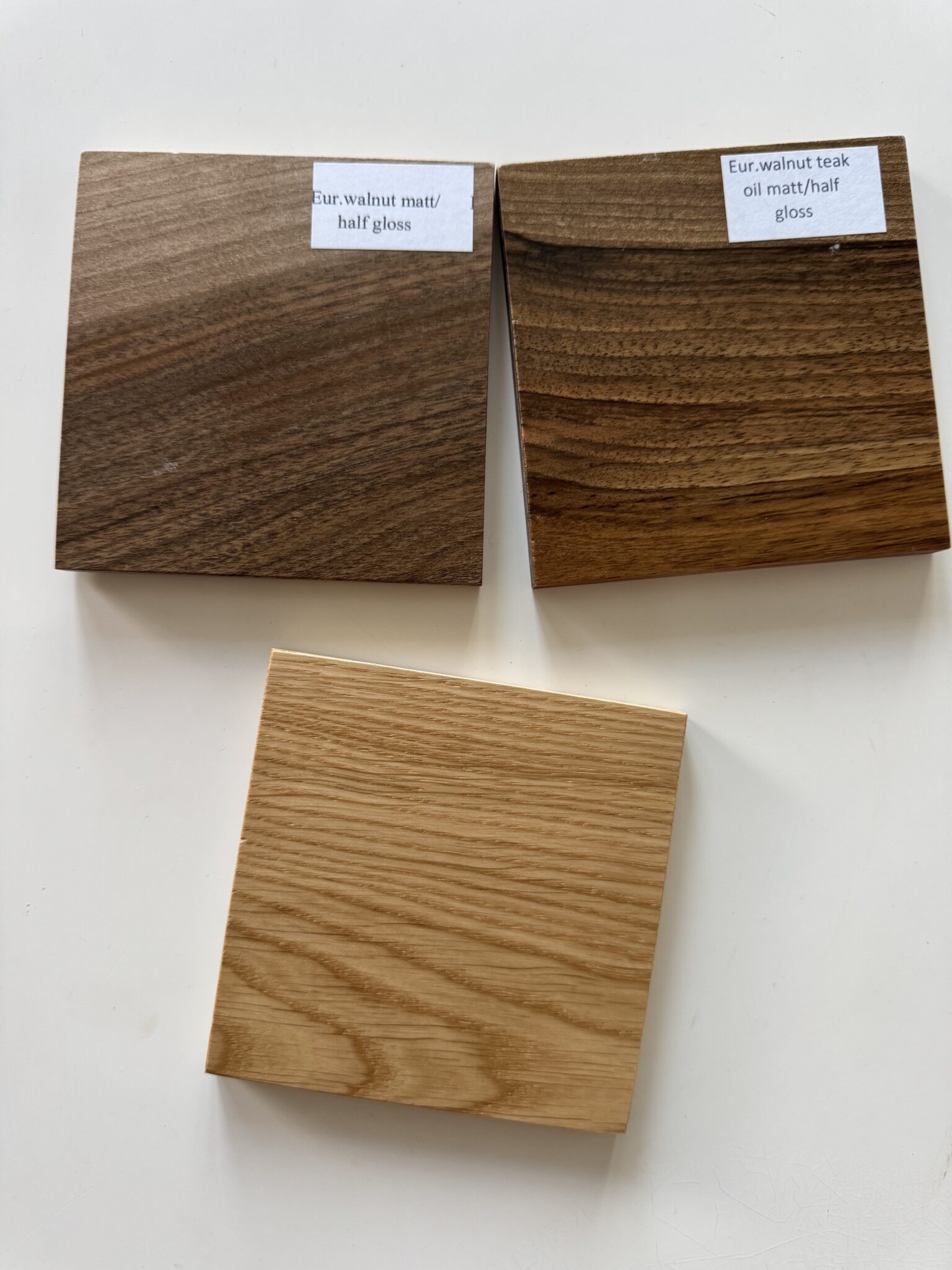 Three wooden samples on a white surface; top left is "Eur. walnut matt/half gloss," top right is "Eur. walnut teak oil matt/half gloss," and bottom is a lighter wood without a label.