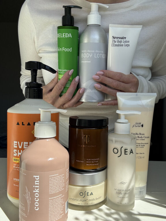 A person holds several bottles of body lotion, with additional skincare and body care products displayed on a table in front of them, all in various shapes and sizes.