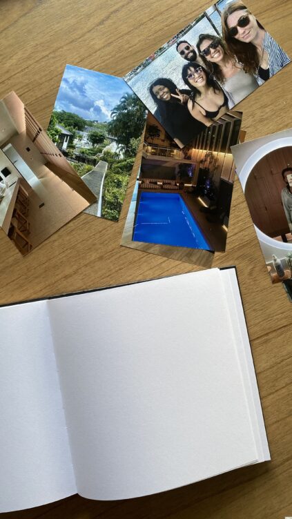 A blank photo album lies open on a wooden table next to several printed photos, including group selfies and images of buildings, a pool, and landscapes.