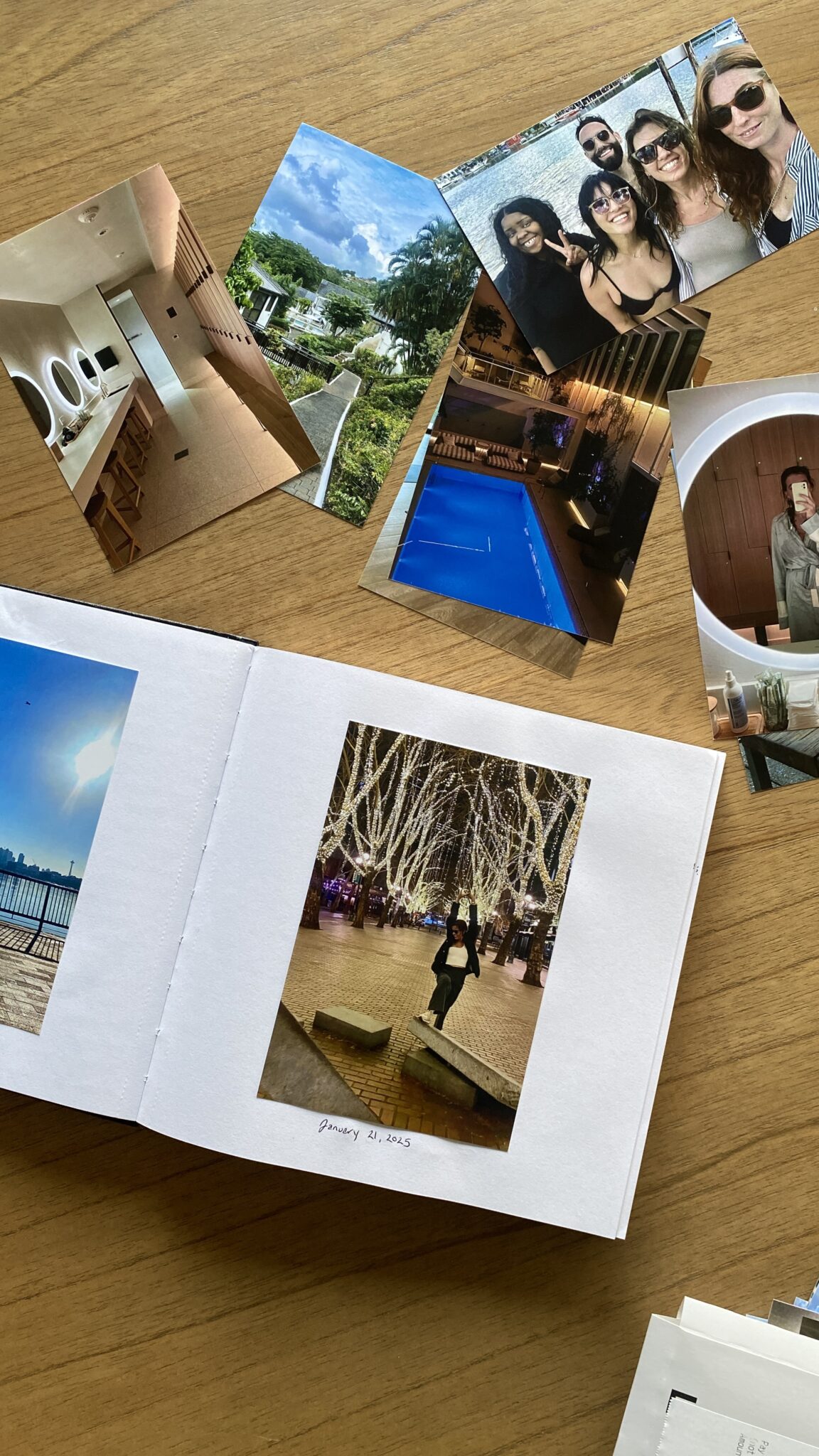 Several printed photos and an open photo book are spread out on a wooden table, showing various scenes including people, buildings, and outdoor locations.