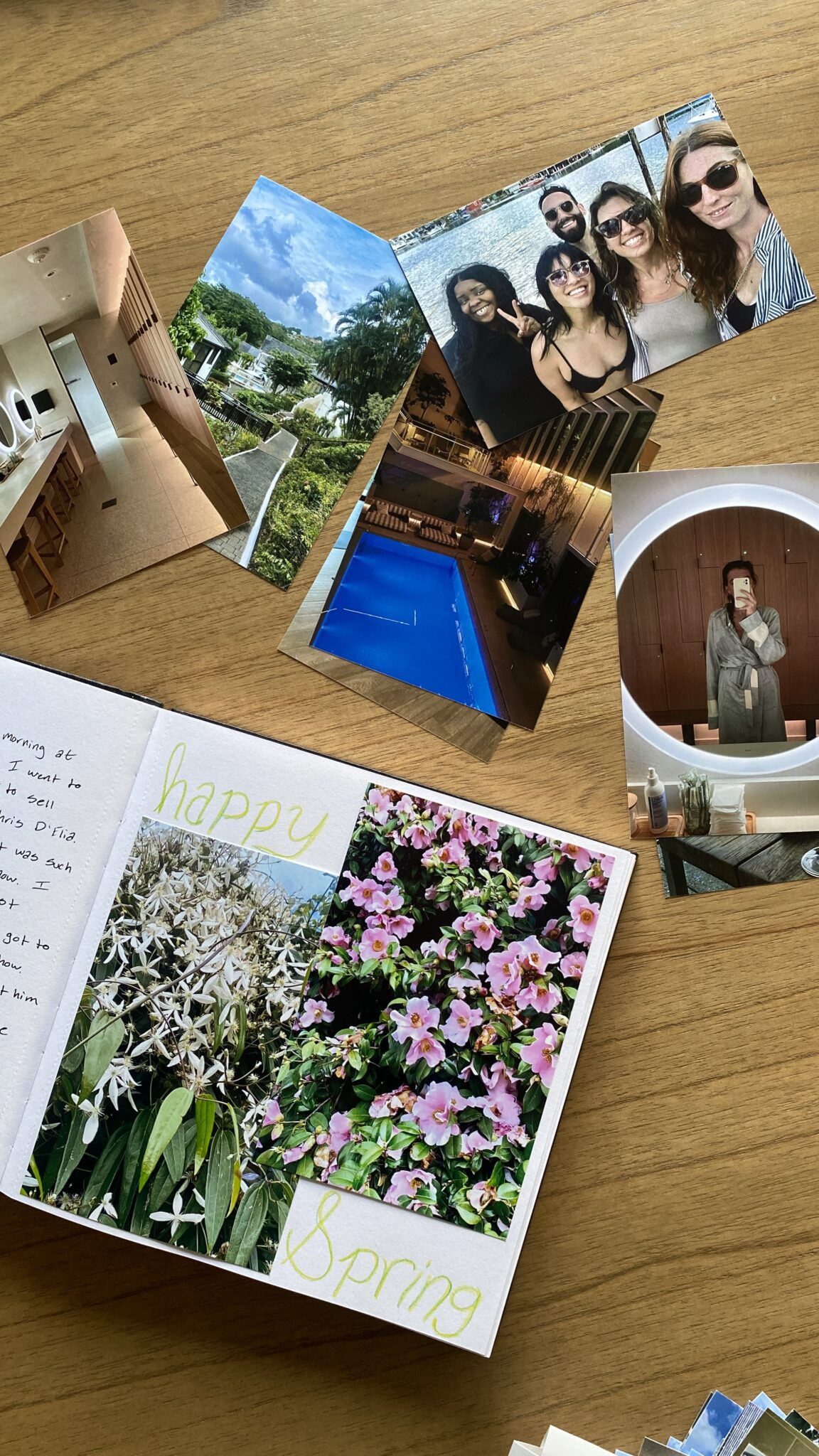 Several printed photos, a journal with "happy Spring" written inside, and a pen are arranged on a wooden table.