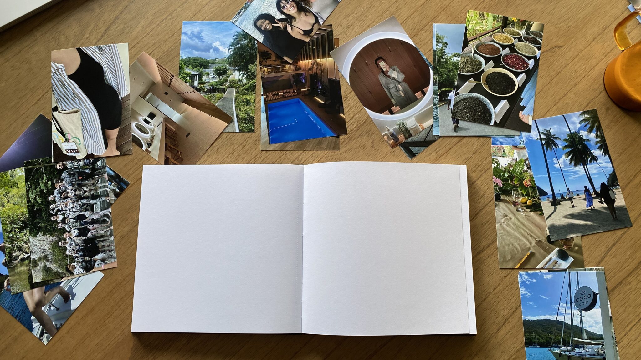 A blank open scrapbook is on a wooden table, surrounded by various printed travel and personal photographs arranged in a semicircle.