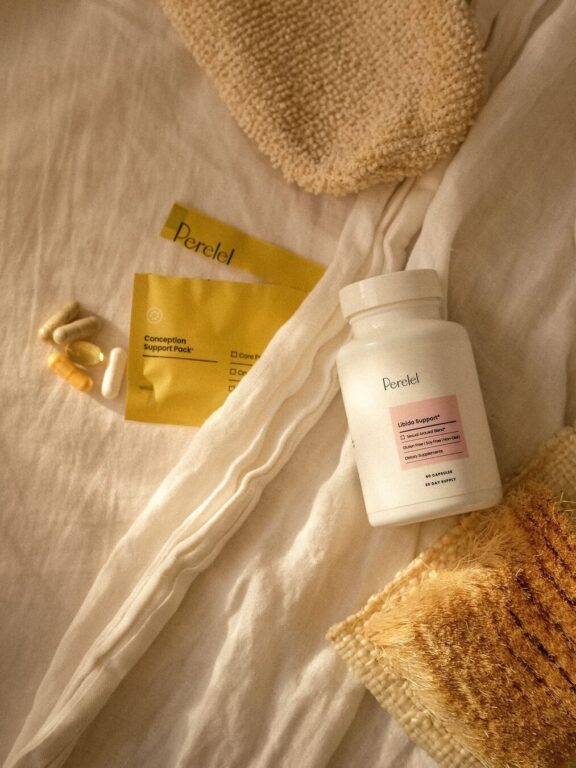 A white bottle labeled "Peretel" sits on a beige fabric next to a yellow packet and several capsules and softgels.