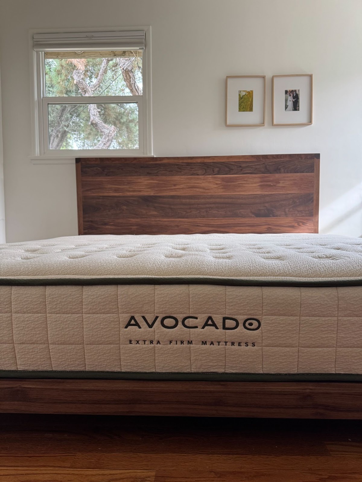A neatly made bed with an Avocado extra firm mattress on a wooden bed frame, set against a white wall with two small framed pictures and a window.