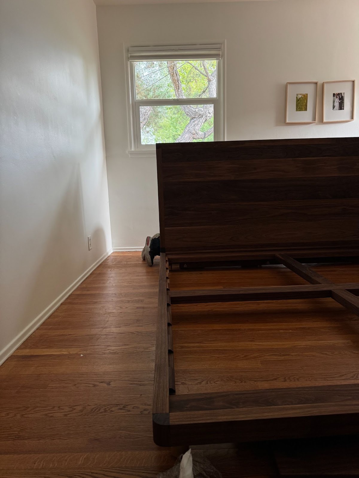 A wooden bed frame sits on a hardwood floor in a minimally furnished room with white walls, a window, and two framed pictures on the wall.