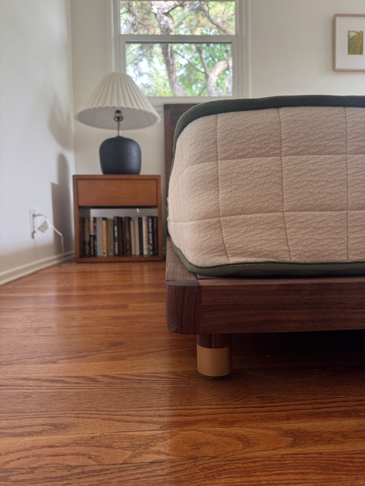 A modern bedroom with a wooden bed frame, a beige mattress, a nightstand with books, a black lamp, and a window showing tree branches.
